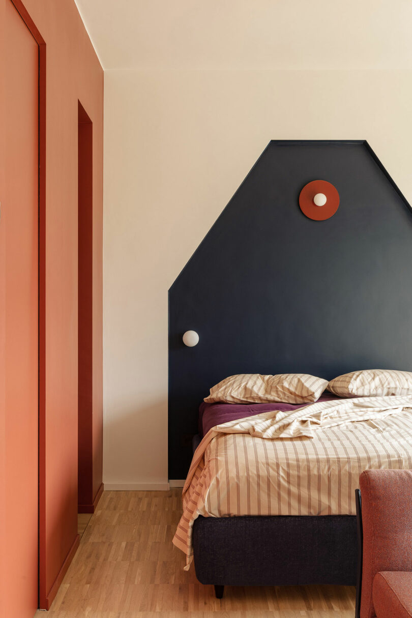A modern bedroom with a bed against a navy geometric accent wall, striped bedding, and orange-red walls and decor accents. Light wood floor.