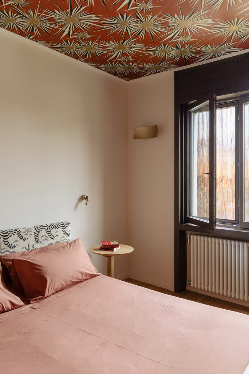 A bedroom with a pink bed, patterned headboard, small round side table, window with black trim, and a ceiling featuring a bold geometric pattern.