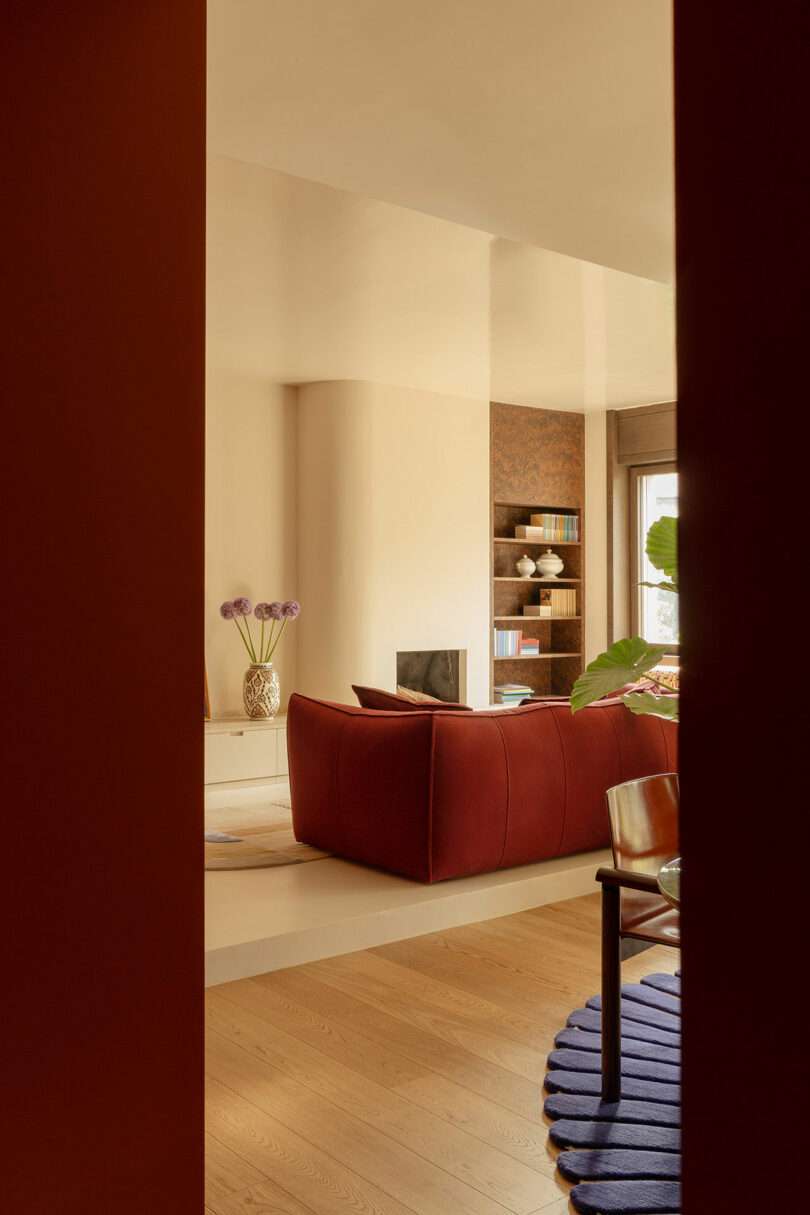 A modern living room with a red sofa, a vase with purple flowers, built-in shelves, and wood flooring, viewed through a doorway.