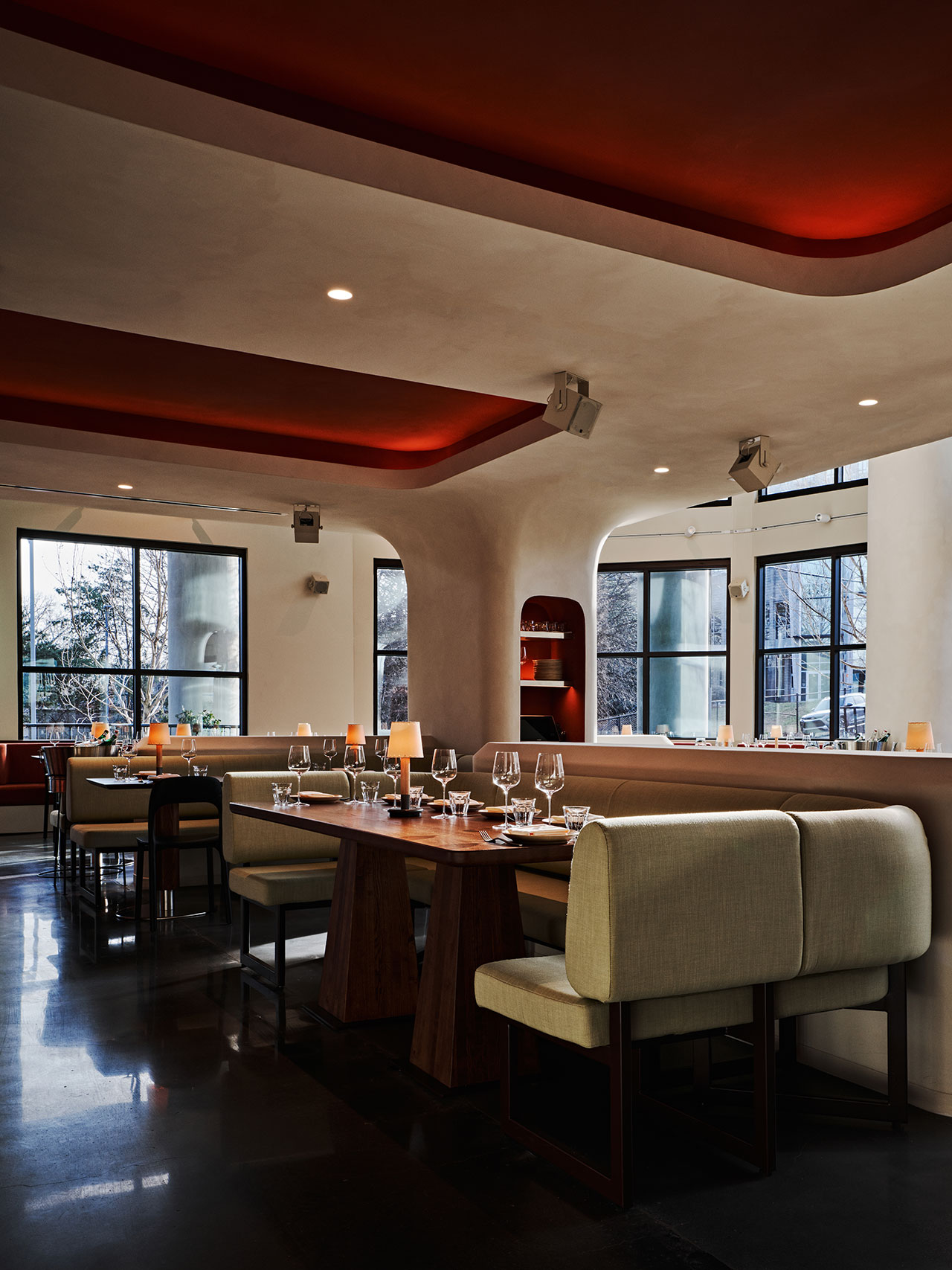 Modern restaurant interior with curved ceiling, red recessed lighting, large windows, beige banquette seating, set tables, and dark reflective flooring.