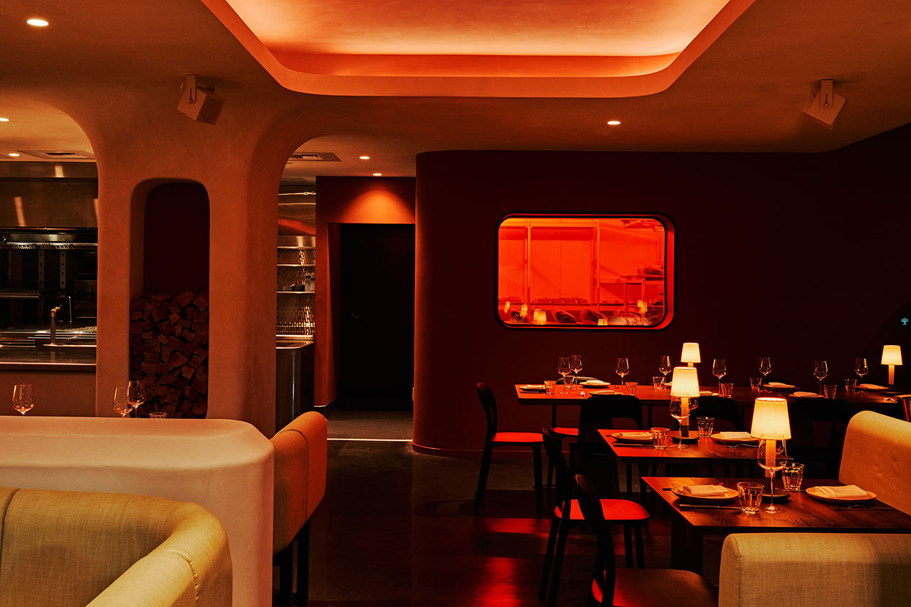 A modern restaurant interior with curved walls, dim lighting, set tables, and an orange-lit window into the kitchen area.