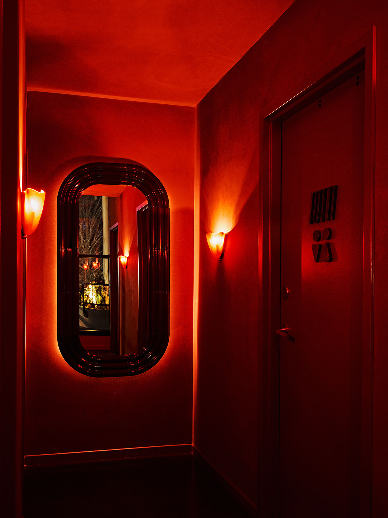 A dimly lit hallway with red walls, a large oval mirror, wall sconces, and a closed door featuring a minimalist restroom sign.