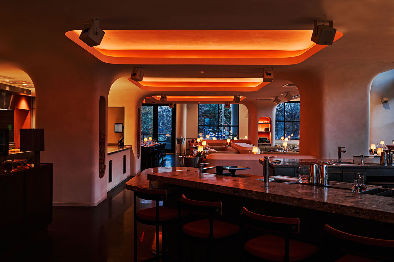 A modern restaurant interior with curved walls, a marble bar counter, dim lighting, and orange-hued ceiling lights, with windows showing trees outside.