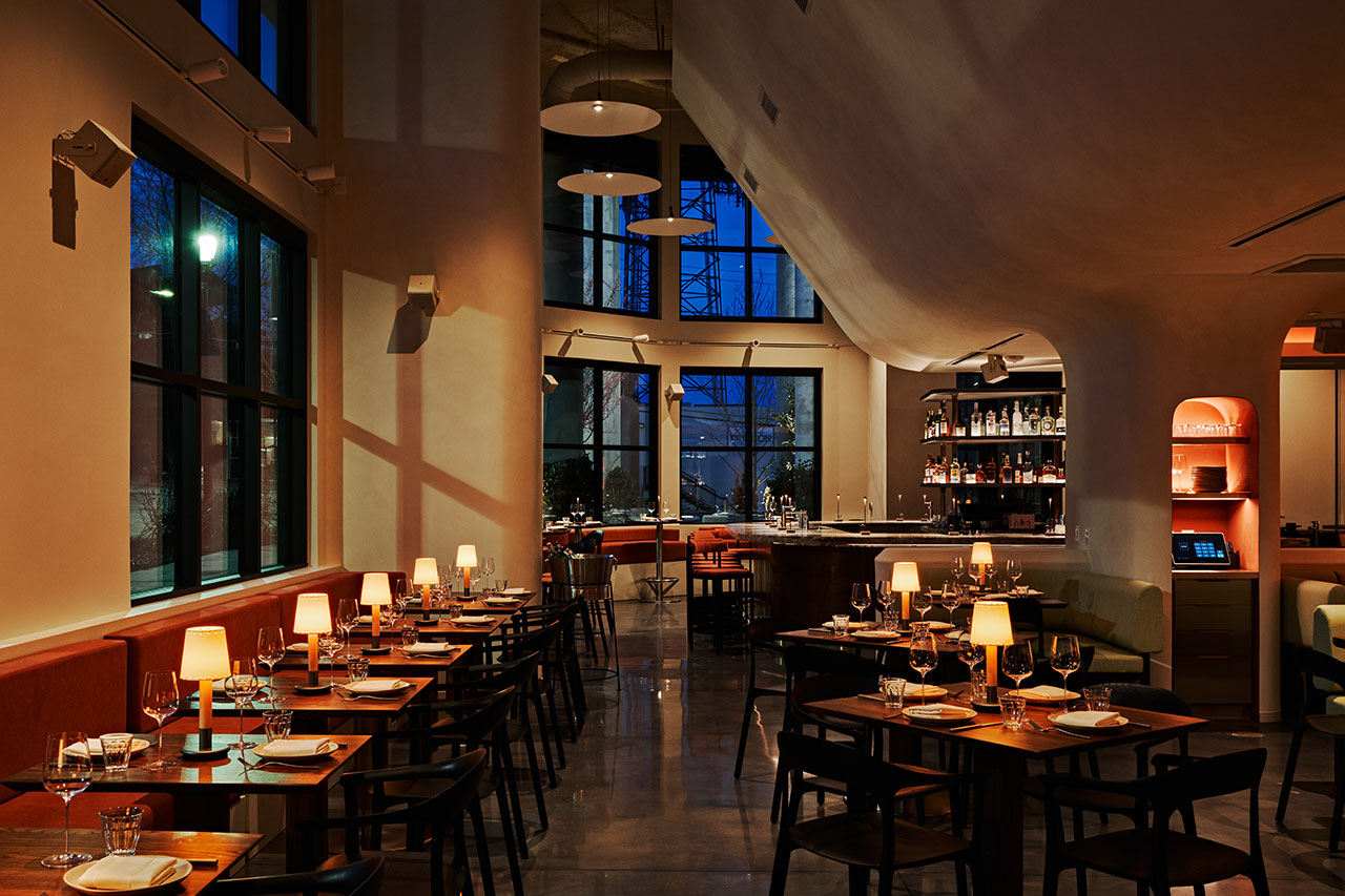Modern restaurant interior with neatly set tables, soft lighting from small table lamps, and a bar area in the background during evening hours.