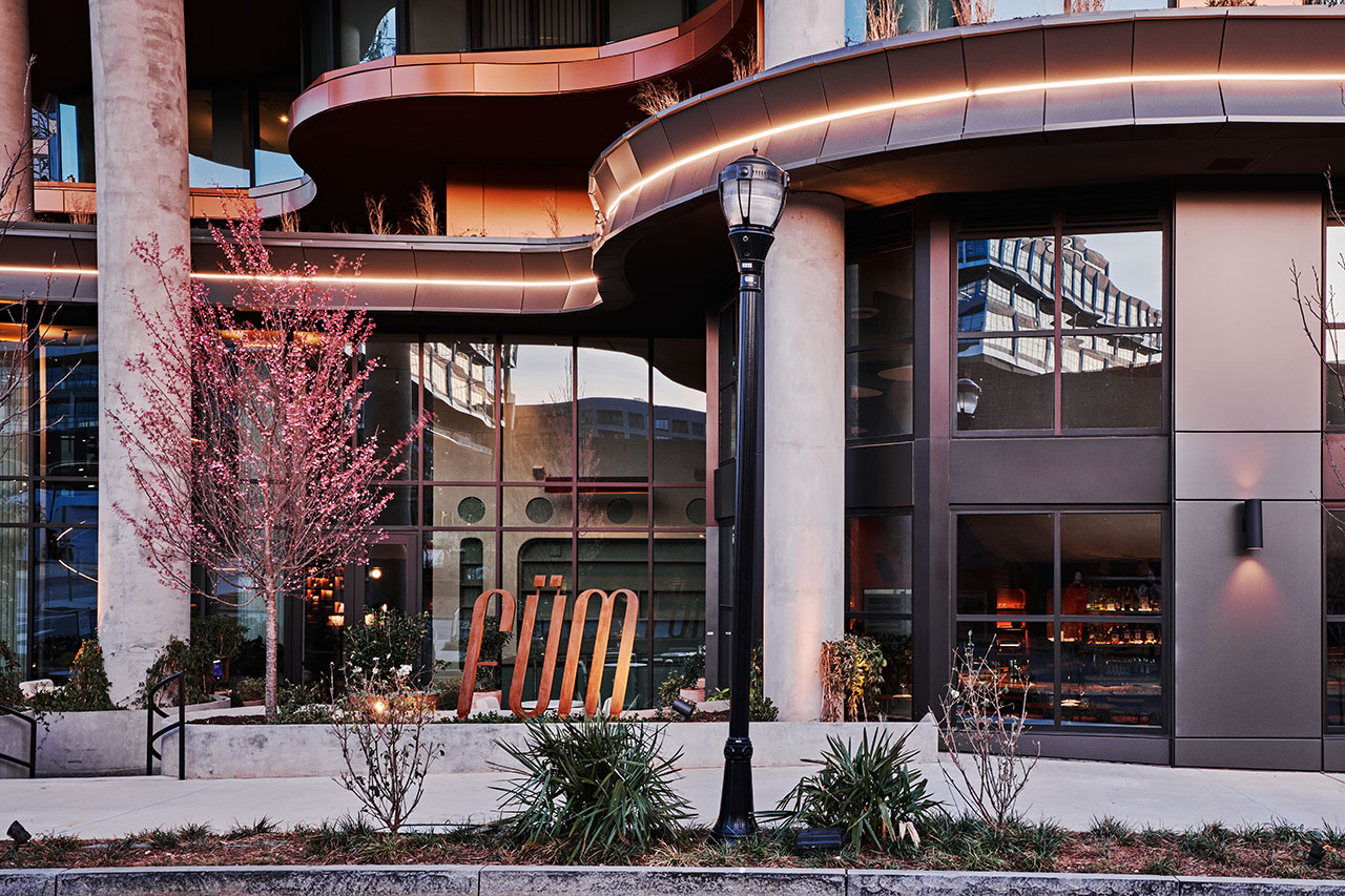 Modern building exterior with large windows, pink-blossomed trees, a streetlamp, and a metal sign reading 