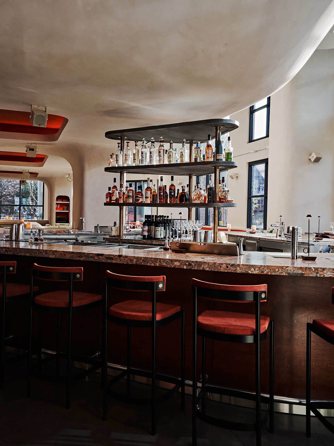 Modern bar with red cushioned stools, marble countertop, and a central liquor display surrounded by glassware under curved, white ceilings and natural light from large windows.