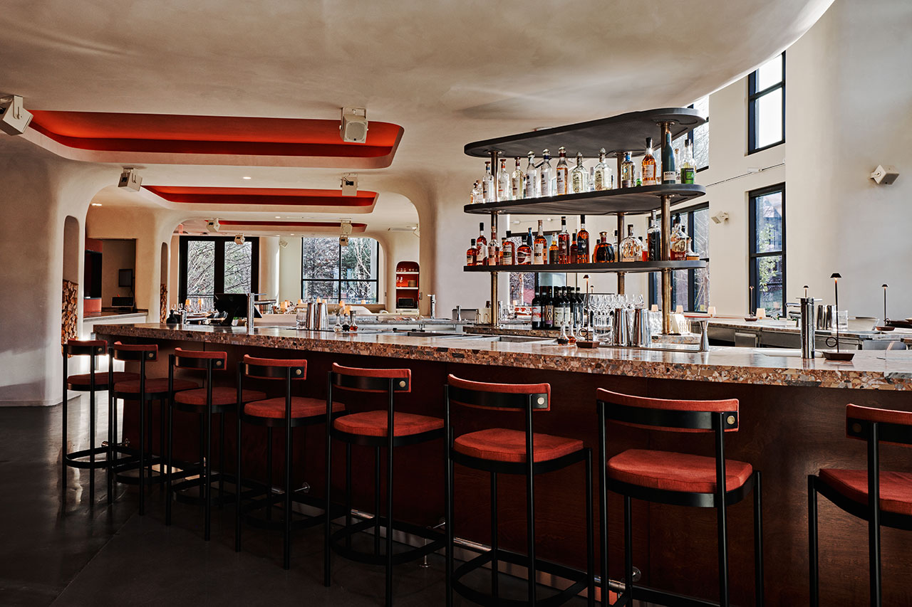 Modern bar with red cushioned stools, marble countertop, and shelves stocked with liquor bottles in a spacious, well-lit interior with curved architectural details.