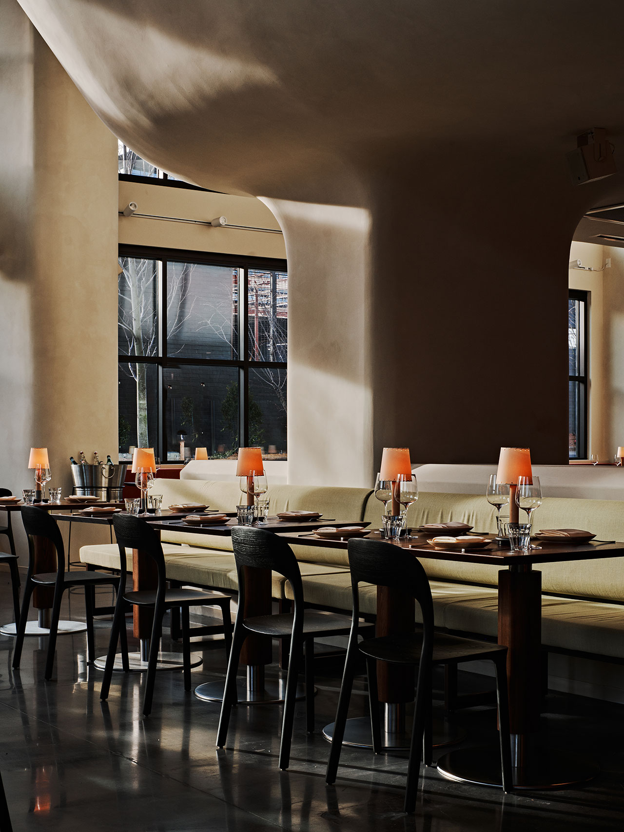 Modern restaurant interior with neatly set tables, soft upholstered seating, orange lamps, and large windows letting in natural light.