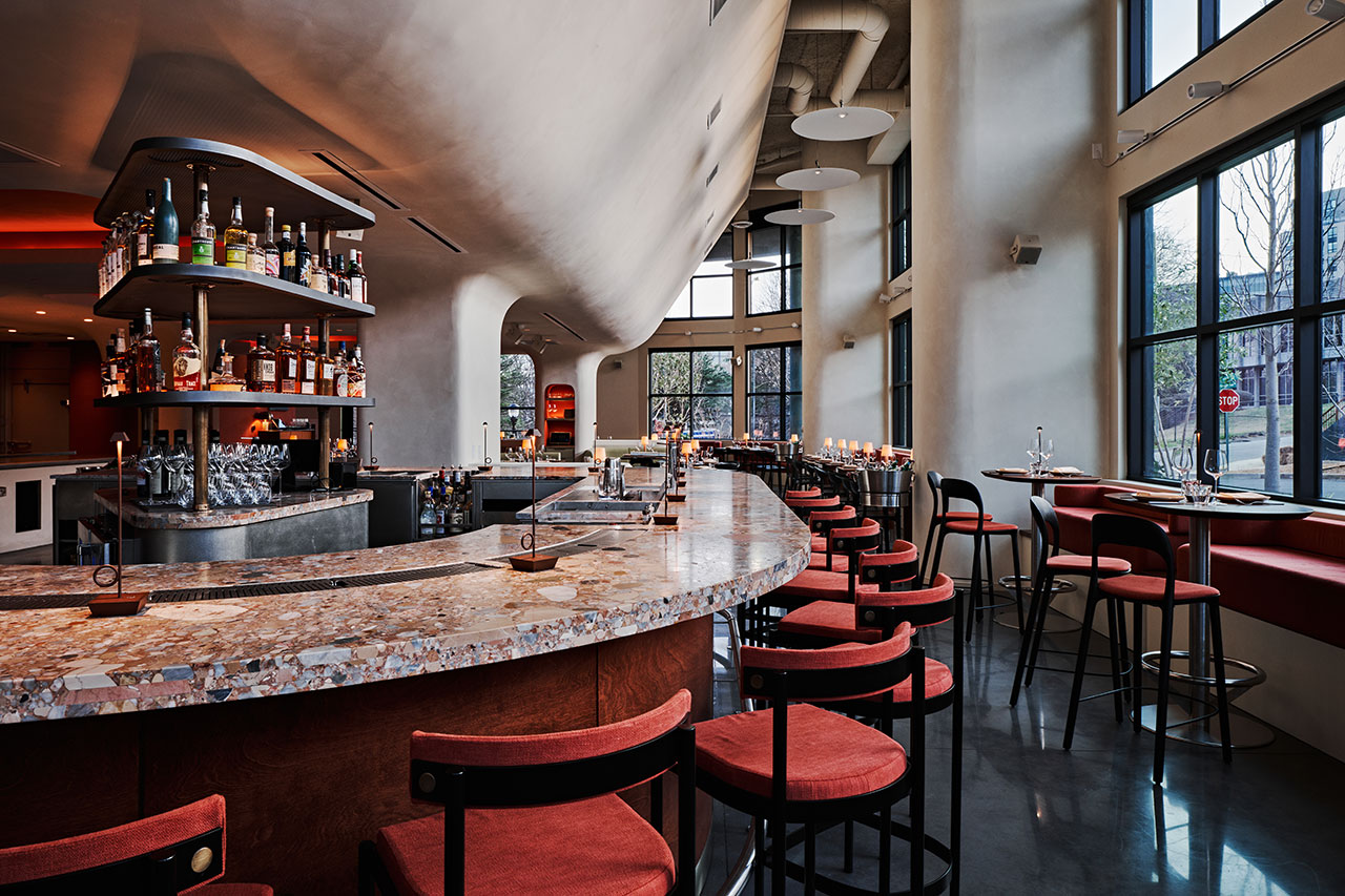 Modern bar interior with a curved marble countertop, red cushioned bar stools, liquor bottles on display, large windows, and natural light filling the space.
