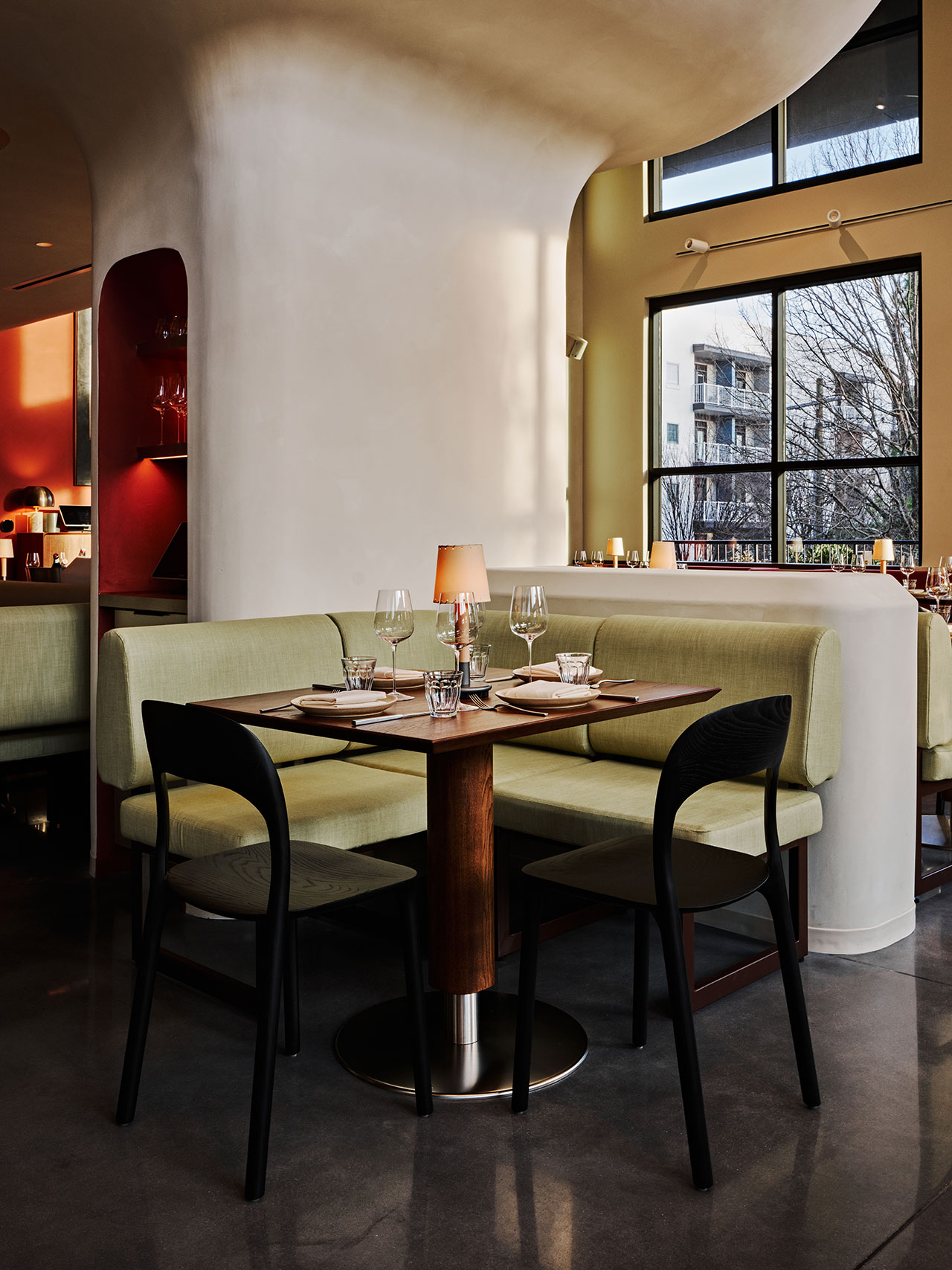 Modern restaurant interior with a set dining table for four, green upholstered booth seating, black chairs, and large windows letting in natural light.