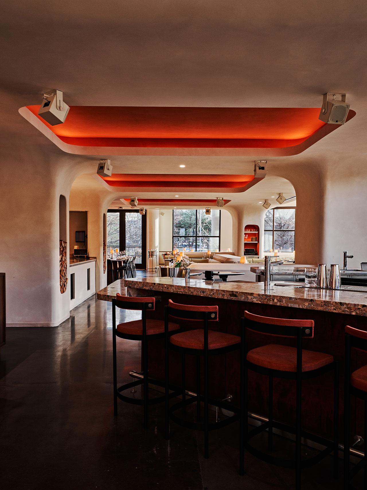 Modern restaurant interior with curved white walls, red accent ceiling, and a bar area with dark wood stools. Large windows in the background let in natural light.