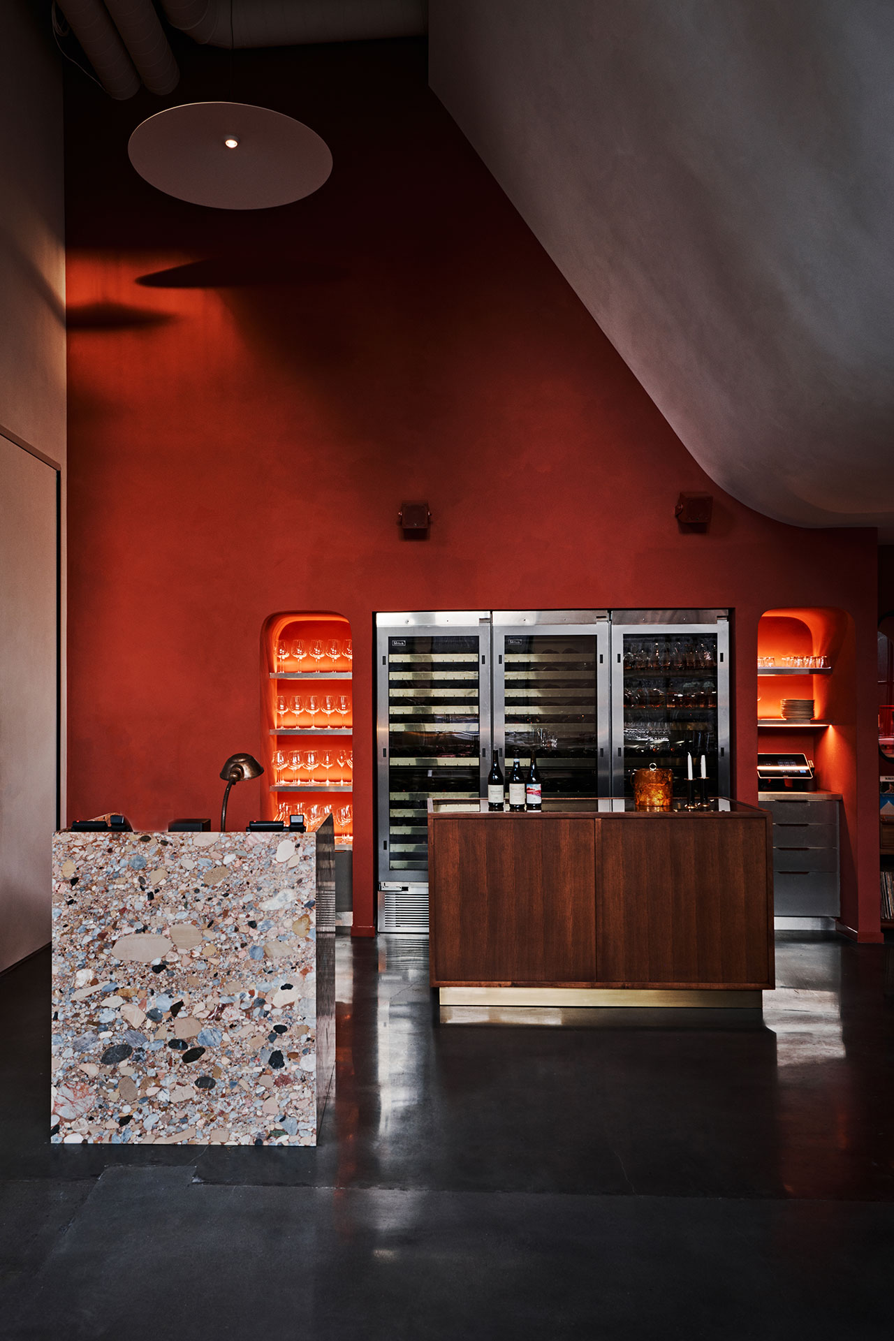 A modern wine bar interior with a terrazzo counter, wooden island, glassware shelves with orange lighting, and stainless steel wine fridges against a red wall.