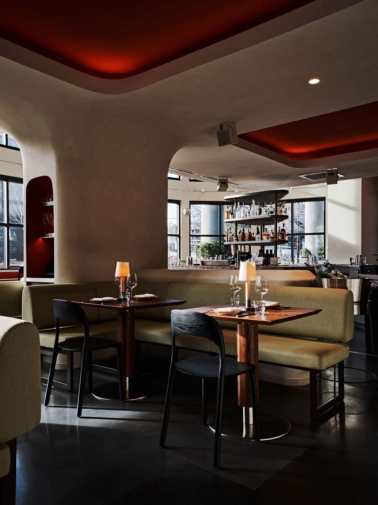 Modern restaurant interior with curved beige booths, dark chairs, set tables with glassware, and large windows letting in natural light.