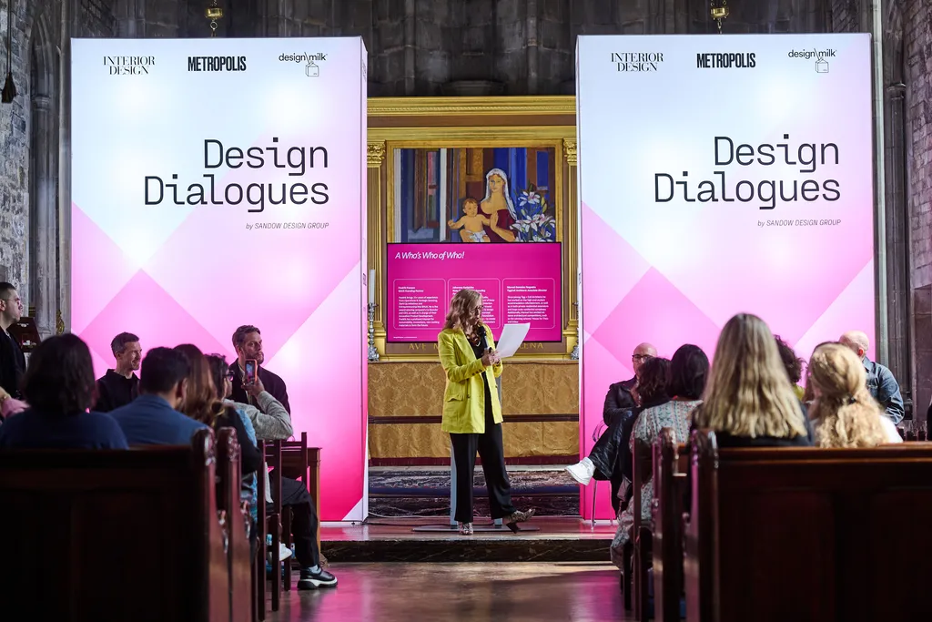 A speaker in a yellow jacket addresses an audience at the "Design Dialogues" event, with large pink and white banners and a painting displayed in the background.
