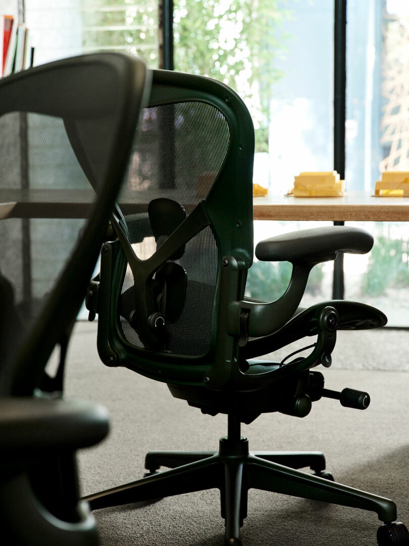 An ergonomic office chair with a mesh backrest sits near a wooden desk in a modern office with large windows and natural light.