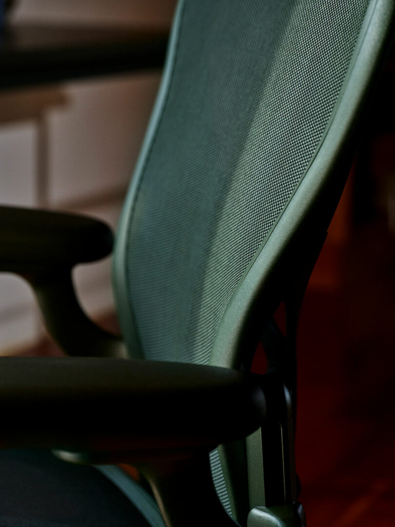 Close-up of a modern ergonomic office chair with a mesh backrest and armrests, shown in a dimly lit indoor setting.