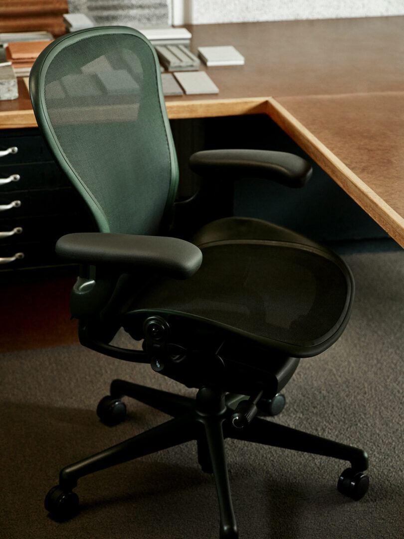 A black ergonomic office chair with mesh back and seat is positioned at the corner of a wooden desk in an office setting.