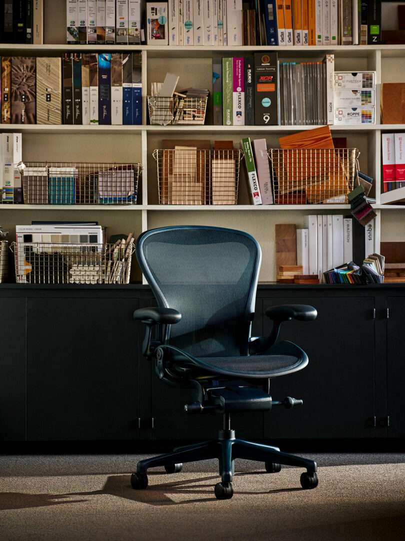 A black ergonomic office chair is positioned in front of black cabinets and shelves filled with books, binders, and wire baskets in a well-lit office space.