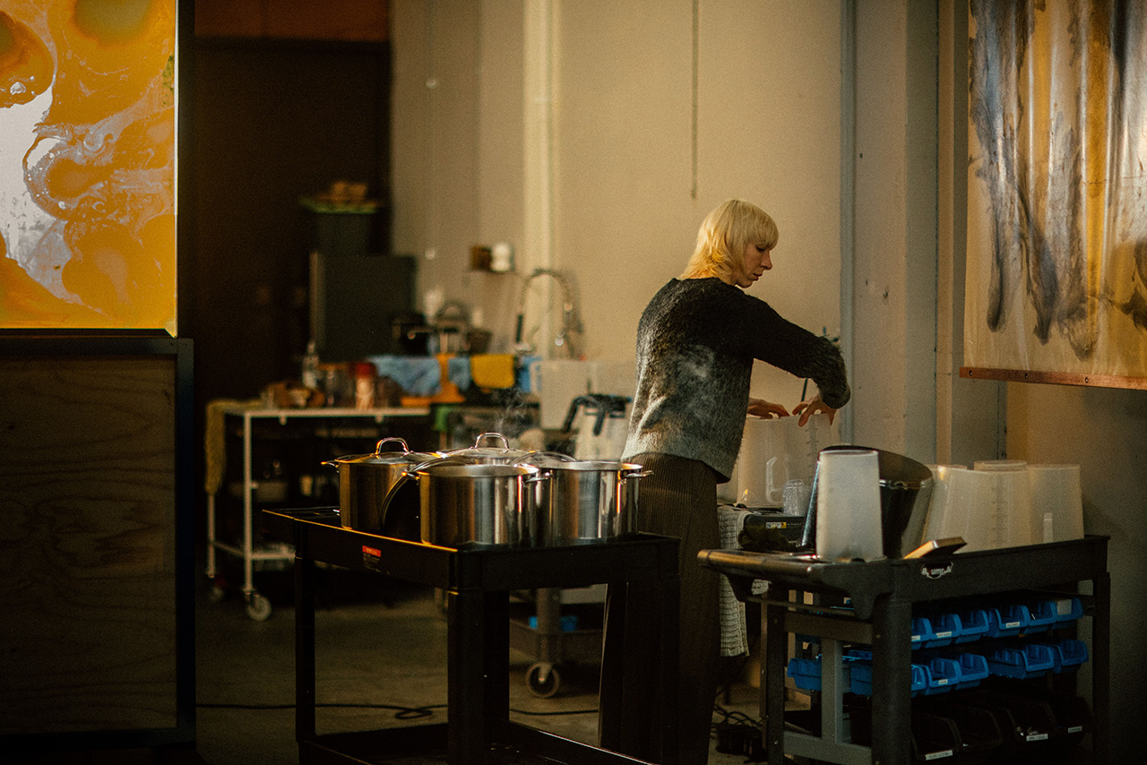 A person with blonde hair stands by a cart with large metal pots in an industrial-style kitchen, evoking the understated elegance of Aesop’s minimalist design.