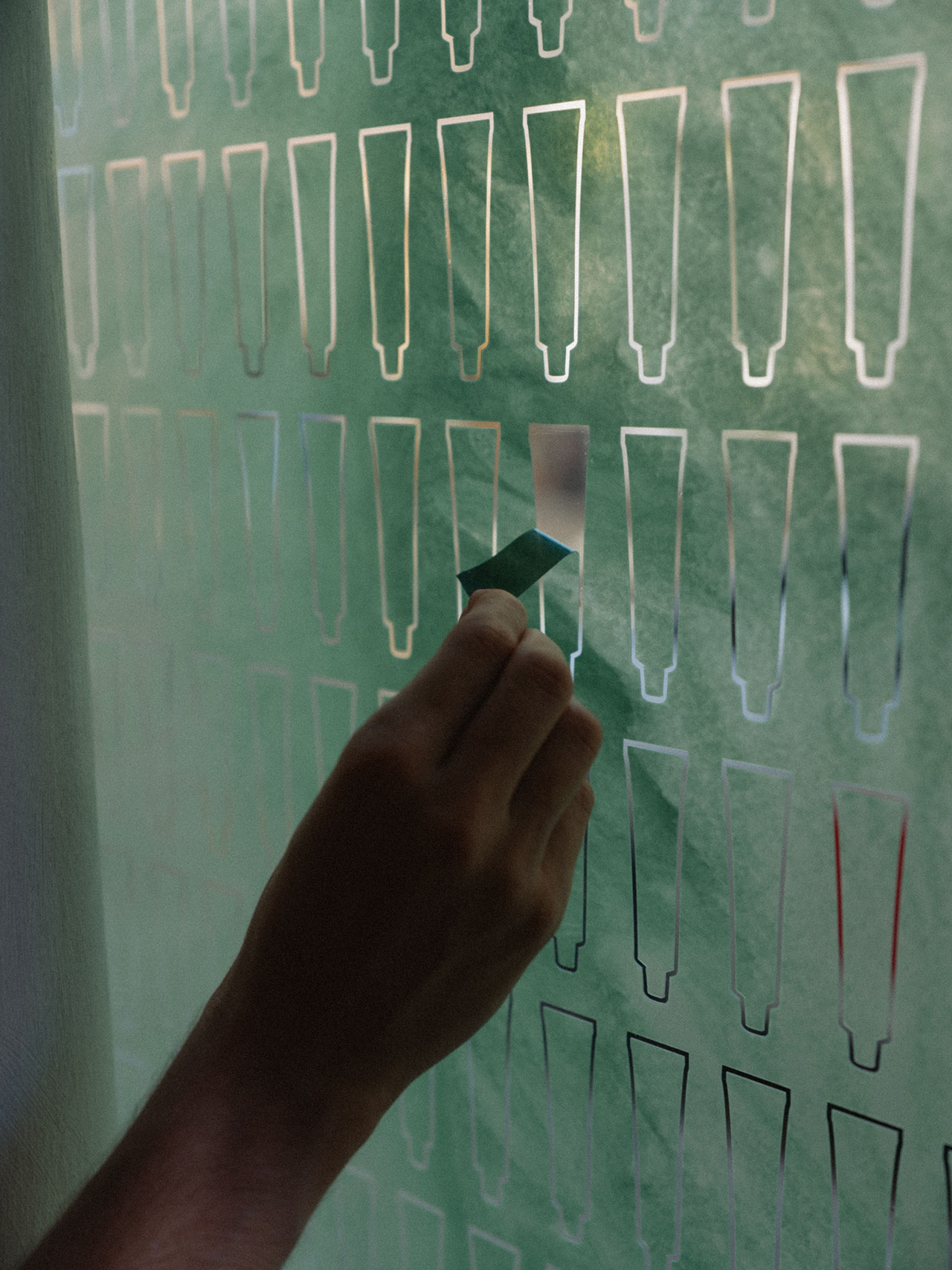 A hand peels a green rectangular sticker from an Aesop stencil sheet with repeated exclamation mark cutouts.