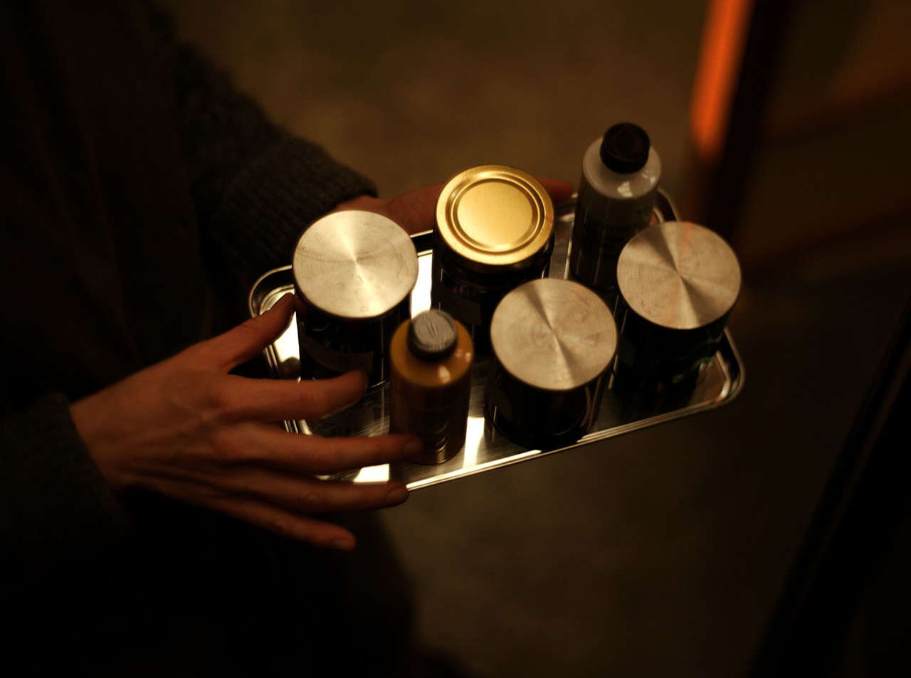 A person holds a metal tray with six variously sized containers, including Aesop jars and bottles, viewed from above in dim lighting.