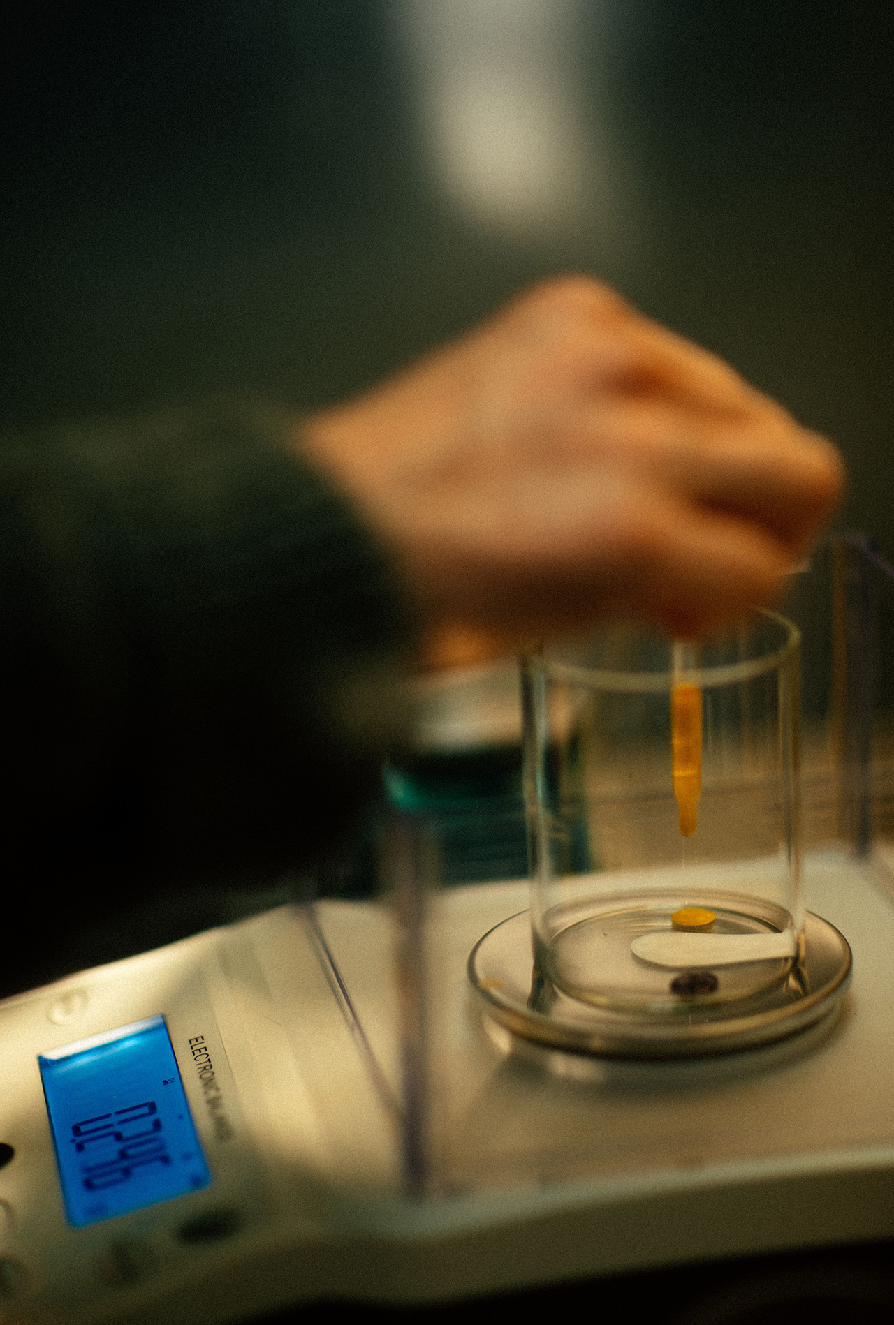 A hand uses a pipette to add liquid into a glass container on an electronic scale, displaying a measurement—a scene reminiscent of Aesop’s meticulous approach to crafting quality.