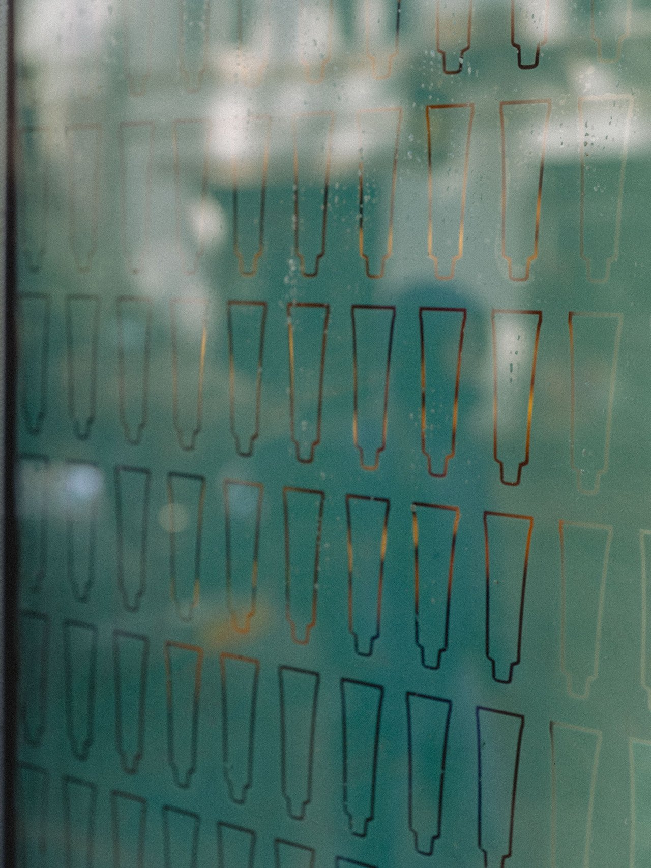A glass panel with a repeated pattern of outlined trapezoidal shapes, slightly fogged with water droplets visible on the surface, evokes the refined simplicity seen in Aesop’s minimalist design aesthetic.