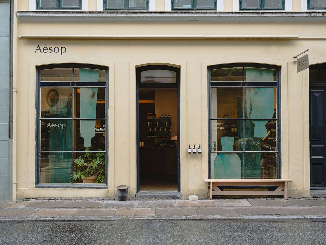 Aesop’s beige storefront features three large arched windows and a door, with neatly displayed shelves of products inside and a wooden bench invitingly set outside.