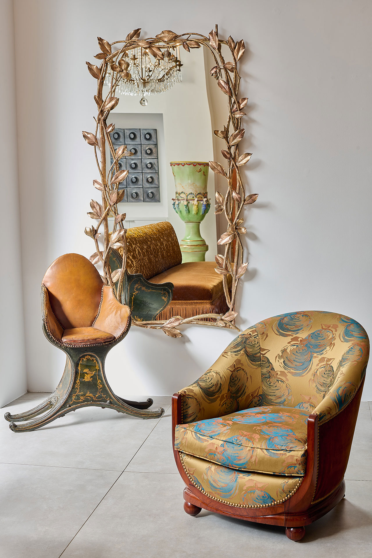 Two vintage chairs sit in front of a large ornate gold mirror with leaf motifs, reflecting a chandelier, wall art, a green vase, and a gold cushion on a sofa.