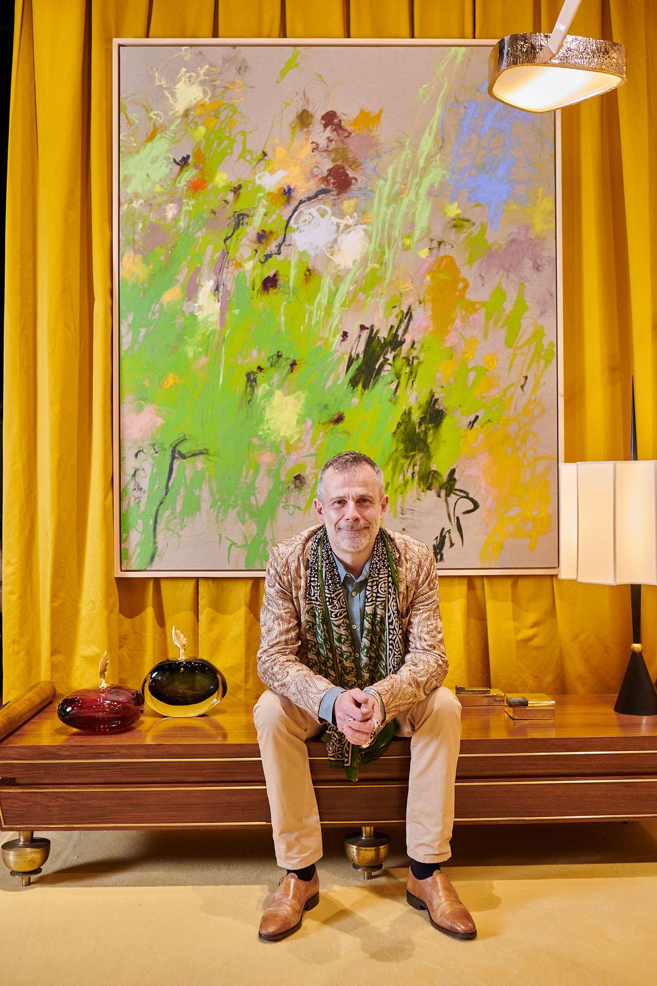 A man sits on a wooden bench in front of a large abstract painting with green and yellow tones, surrounded by modern decor and a yellow curtain background.