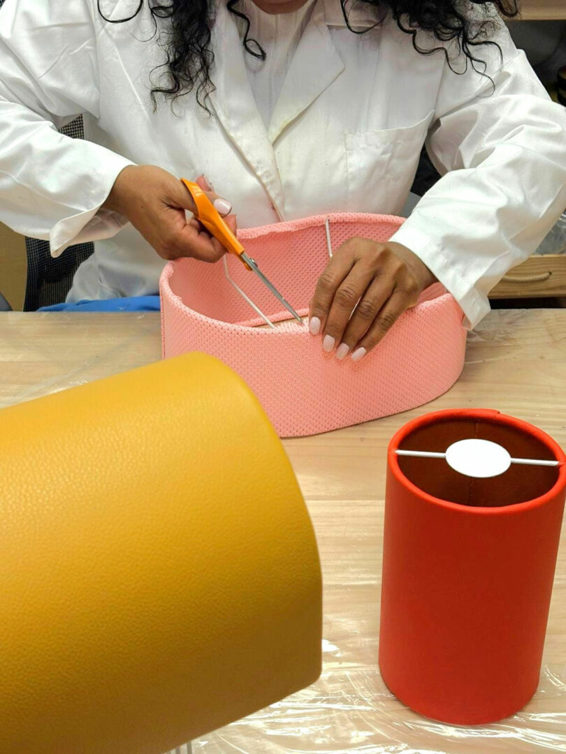 A person in a white lab coat cuts pink fabric with scissors; yellow and red cylindrical objects are on the table.