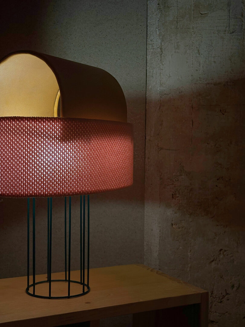 A modern table lamp with a cylindrical shade and metal legs sits on a wooden surface against a concrete wall.