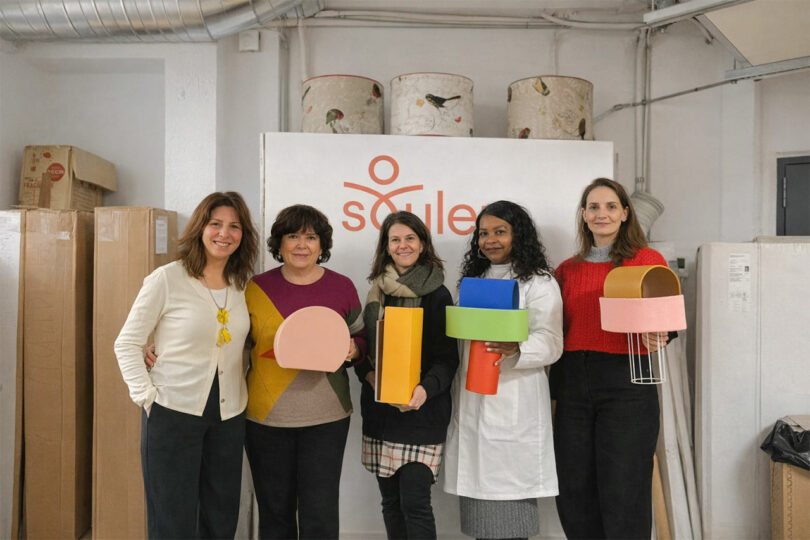 Five women stand indoors in front of a sign reading 