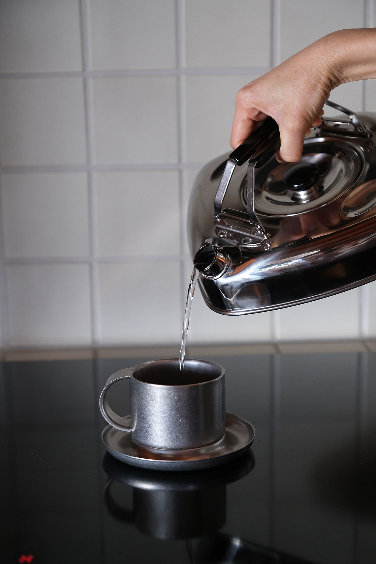 A hand pours hot water from a metal kettle into a gray cup on a saucer, with a tiled wall in the background—a simple building block of daily rituals.