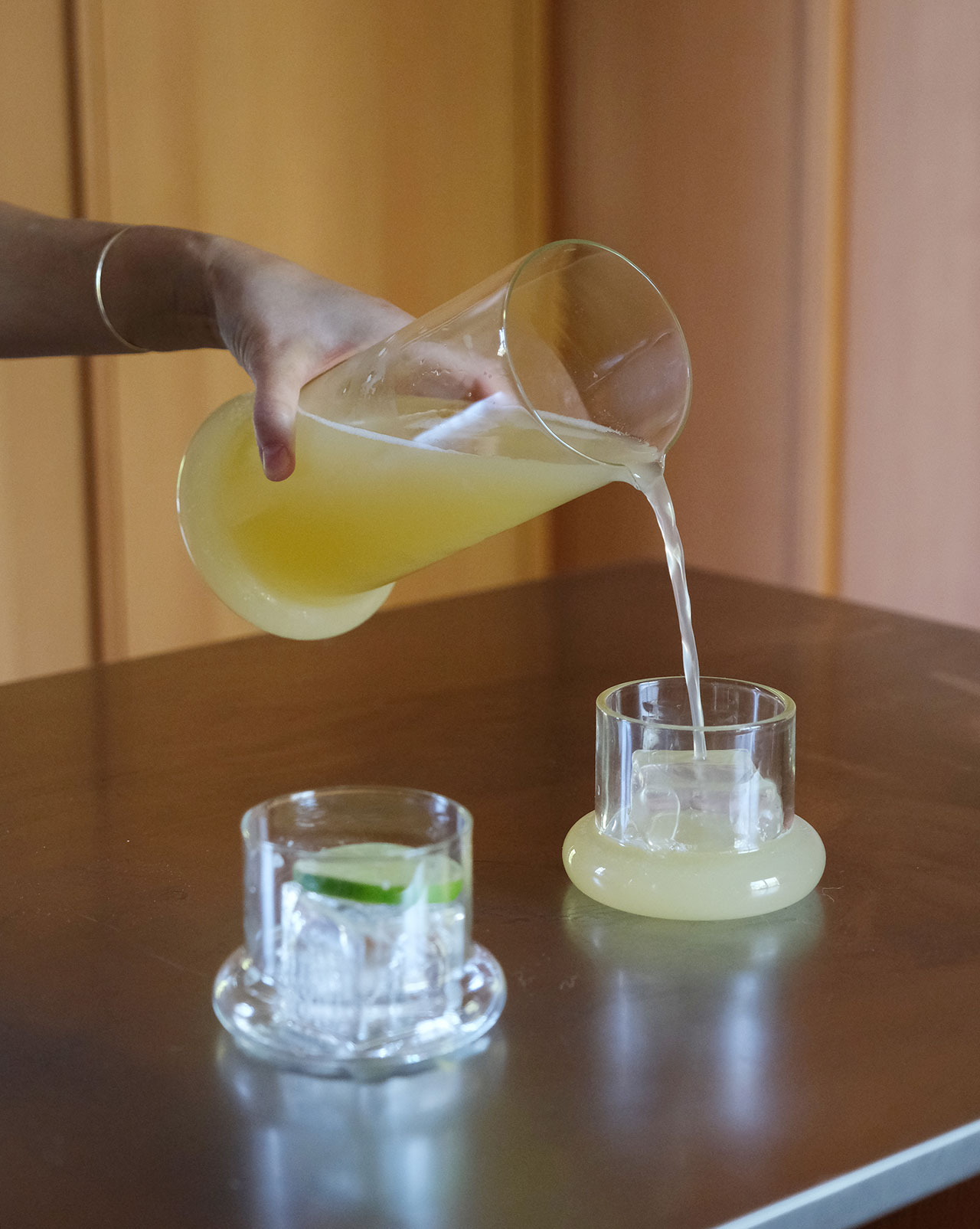 A hand pours yellow juice from a glass pitcher into a glass on a wooden table—a building block of any summer gathering; another glass with ice and a lime wedge sits beside it.