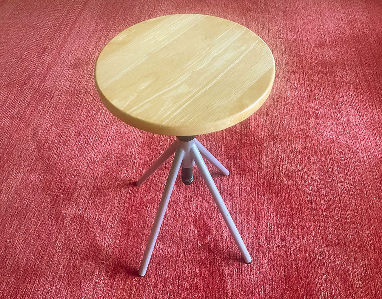 A round wooden stool with three metal legs, designed by Amanda Martocchio, stands on a textured red carpet.