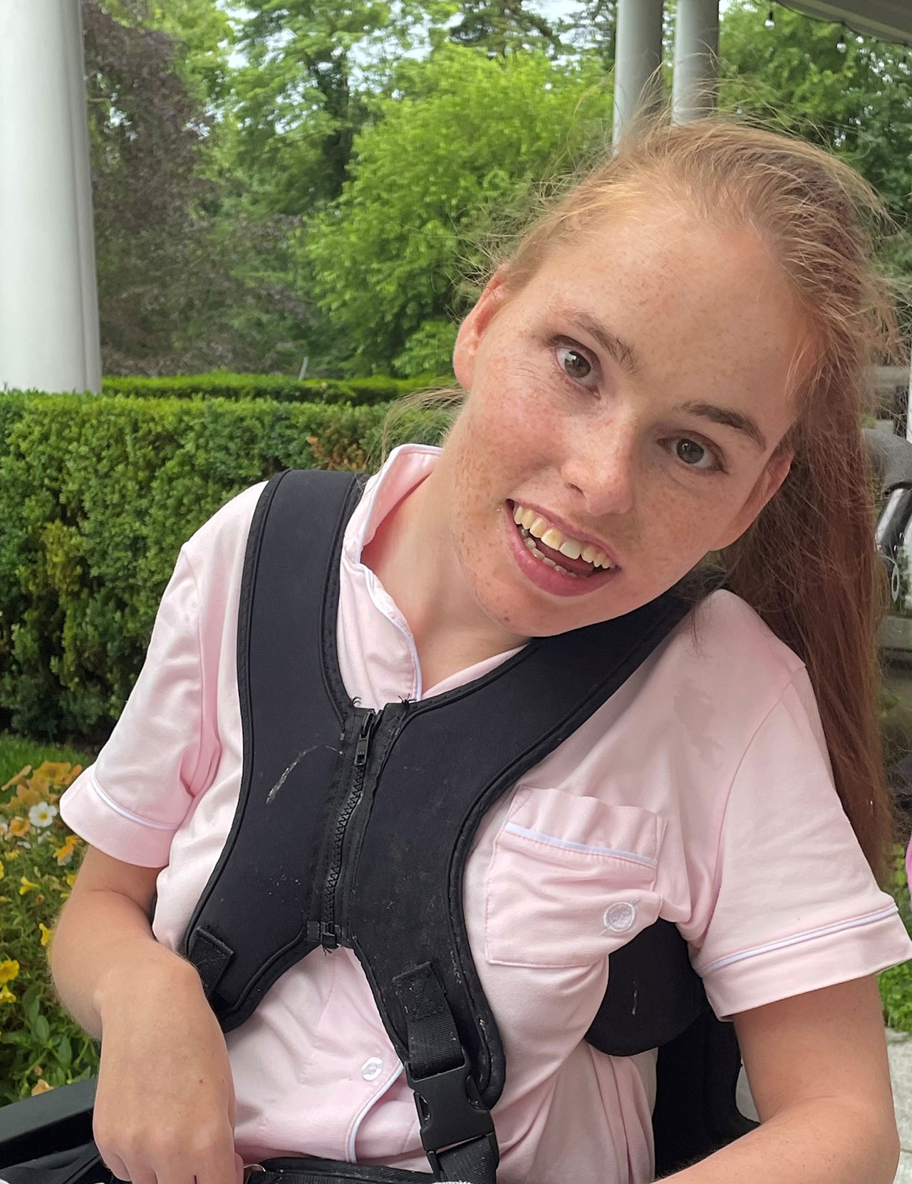 A young woman with long light brown hair, wearing a light pink shirt and a black harness, sits outdoors near green bushes and flowers, smiling at the camera—capturing an inviting scene reminiscent of Amanda Martocchio’s design inspirations.