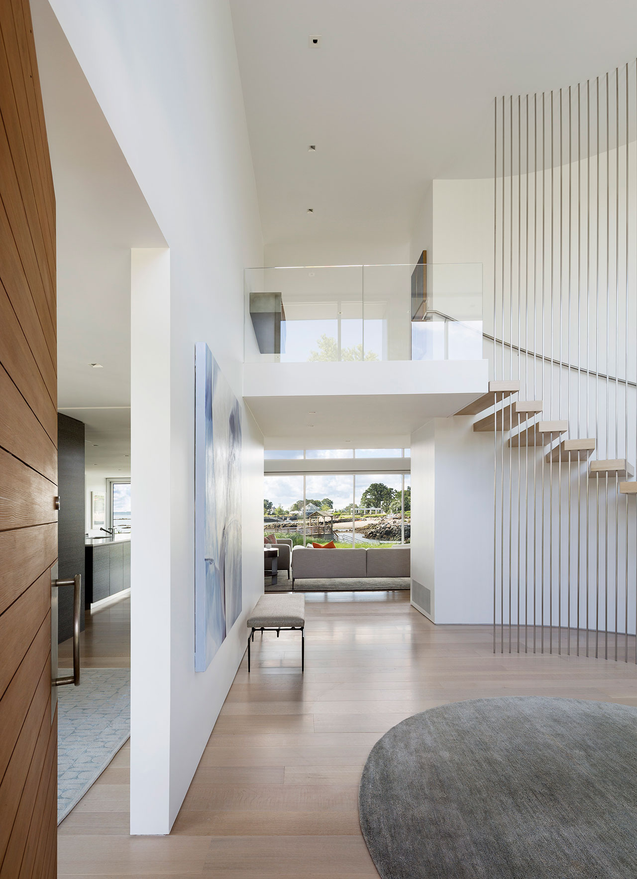 Modern interior by Amanda Martocchio with a floating wooden staircase, glass railing, large windows, light wood floors, white walls, and a gray rug. Artwork and a bench add character to the sunlit hallway.