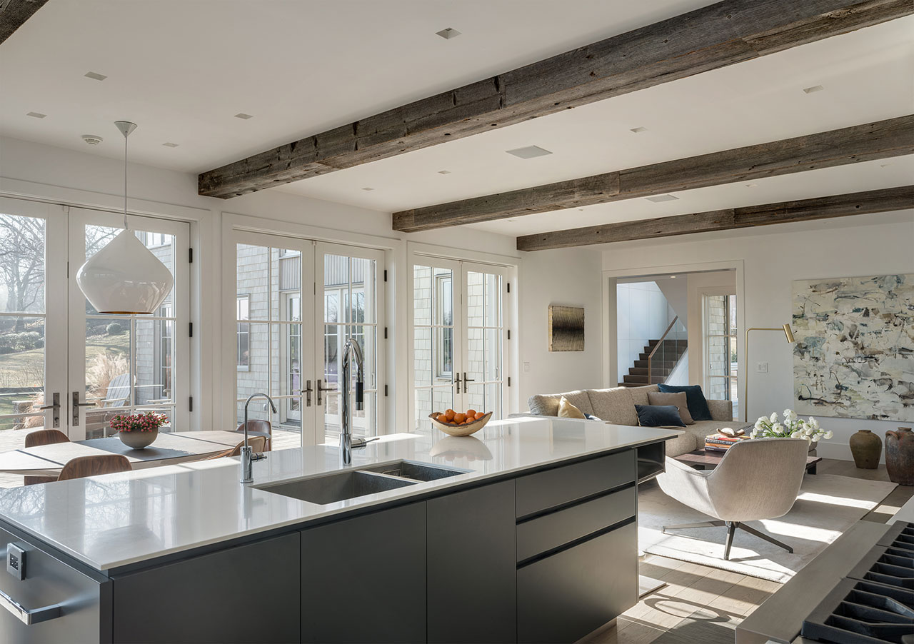 Modern open-concept kitchen and living area designed by Amanda Martocchio, featuring exposed wooden beams, large windows, a central island, and neutral-toned furnishings.
