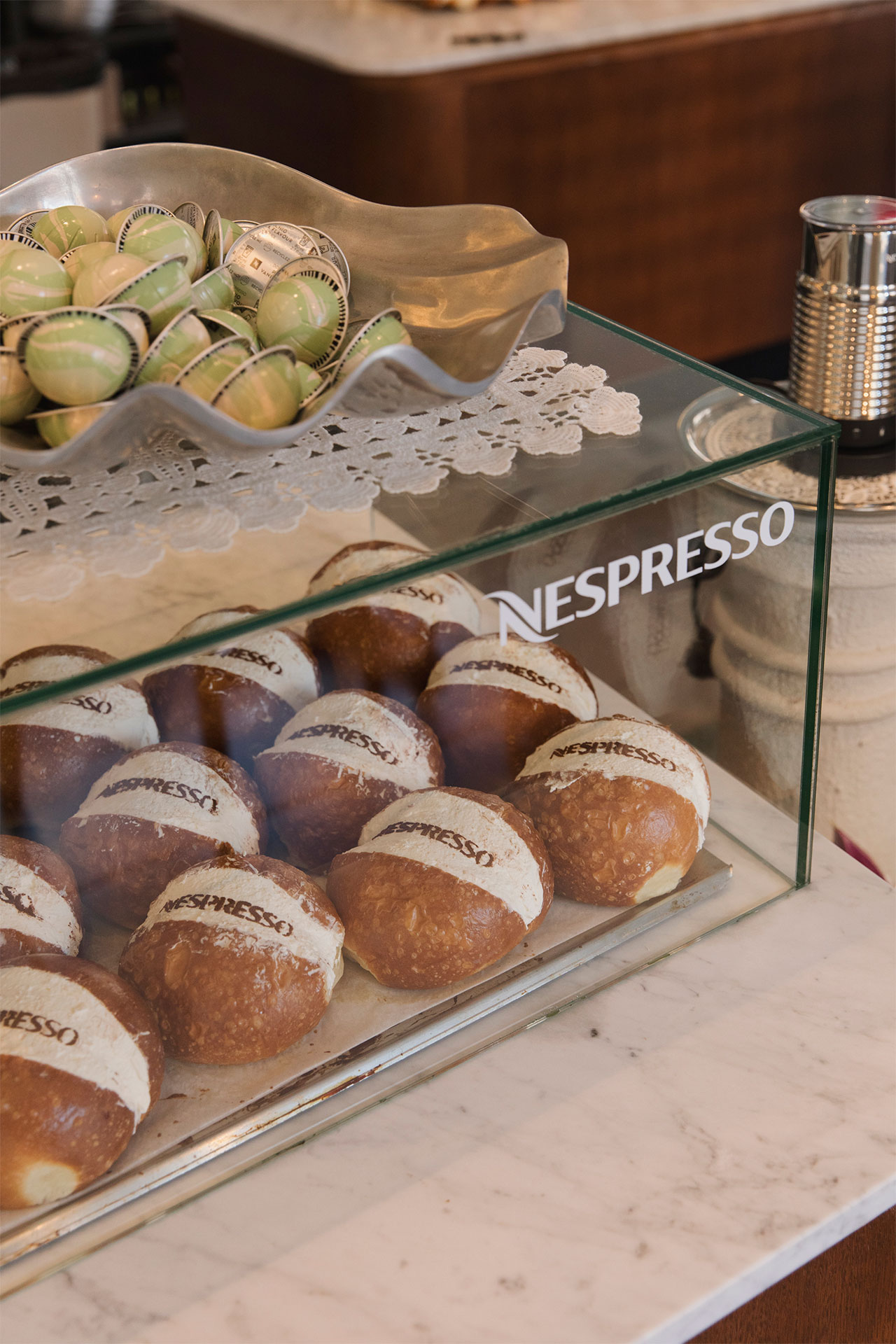 Glass display case with cream-filled pastries labeled “Nespresso” alongside a tray of Nespresso coffee pods, all set on a marble counter.