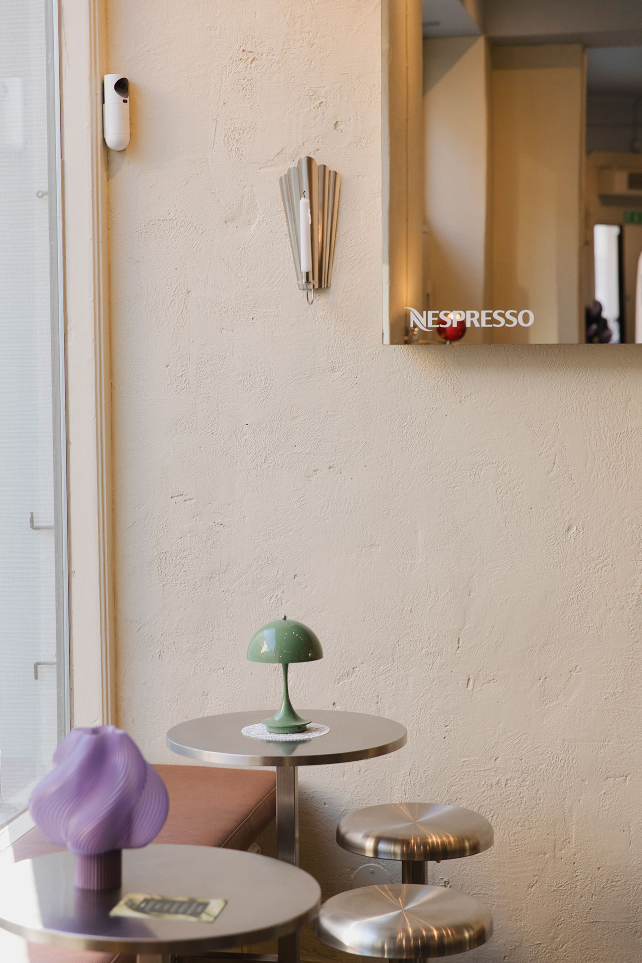Small modern café interior with two round metal tables, a green lamp, a purple decorative object, and cream-colored textured walls. A wall mirror featuring the Nespresso logo adds a stylish touch.