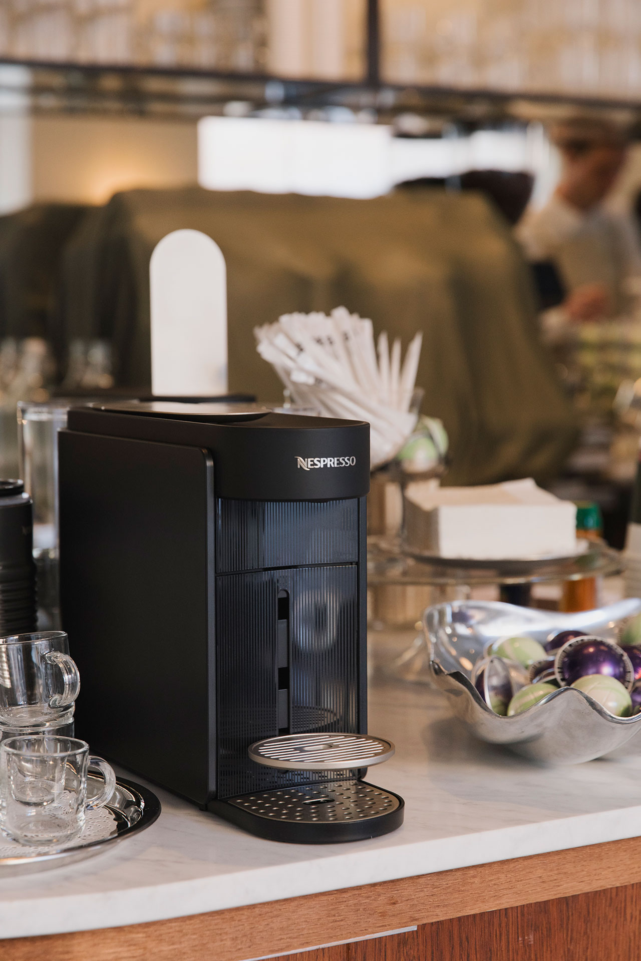 A sleek black Nespresso coffee machine sits on a countertop beside stacked glass cups, a metal bowl of Nespresso pods, and various coffee accessories.