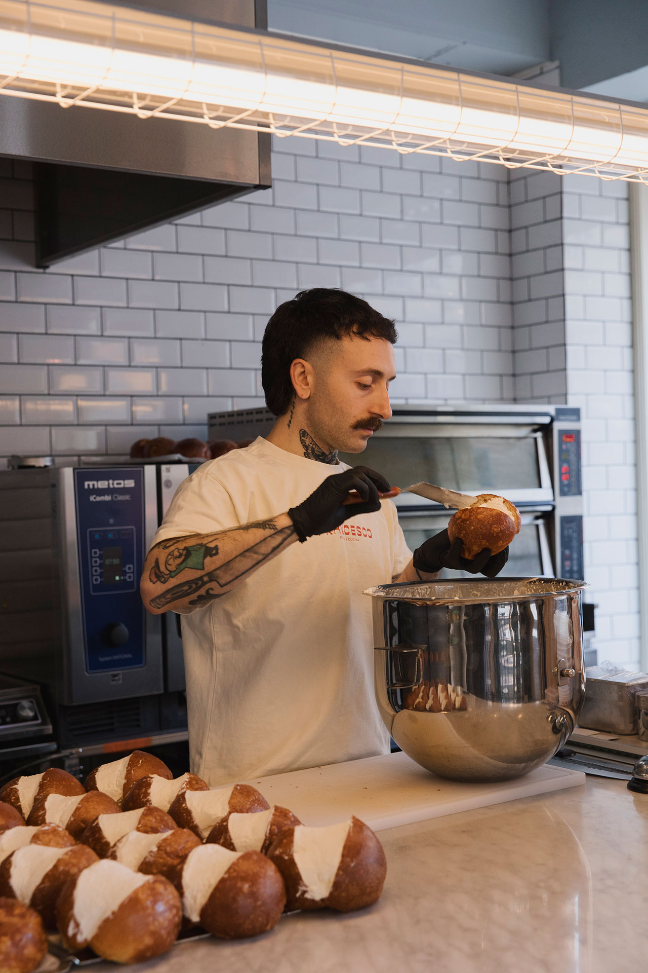 A person wearing gloves spreads cream filling onto a bread roll in a commercial kitchen, with several filled rolls on the counter beside a Nespresso machine.