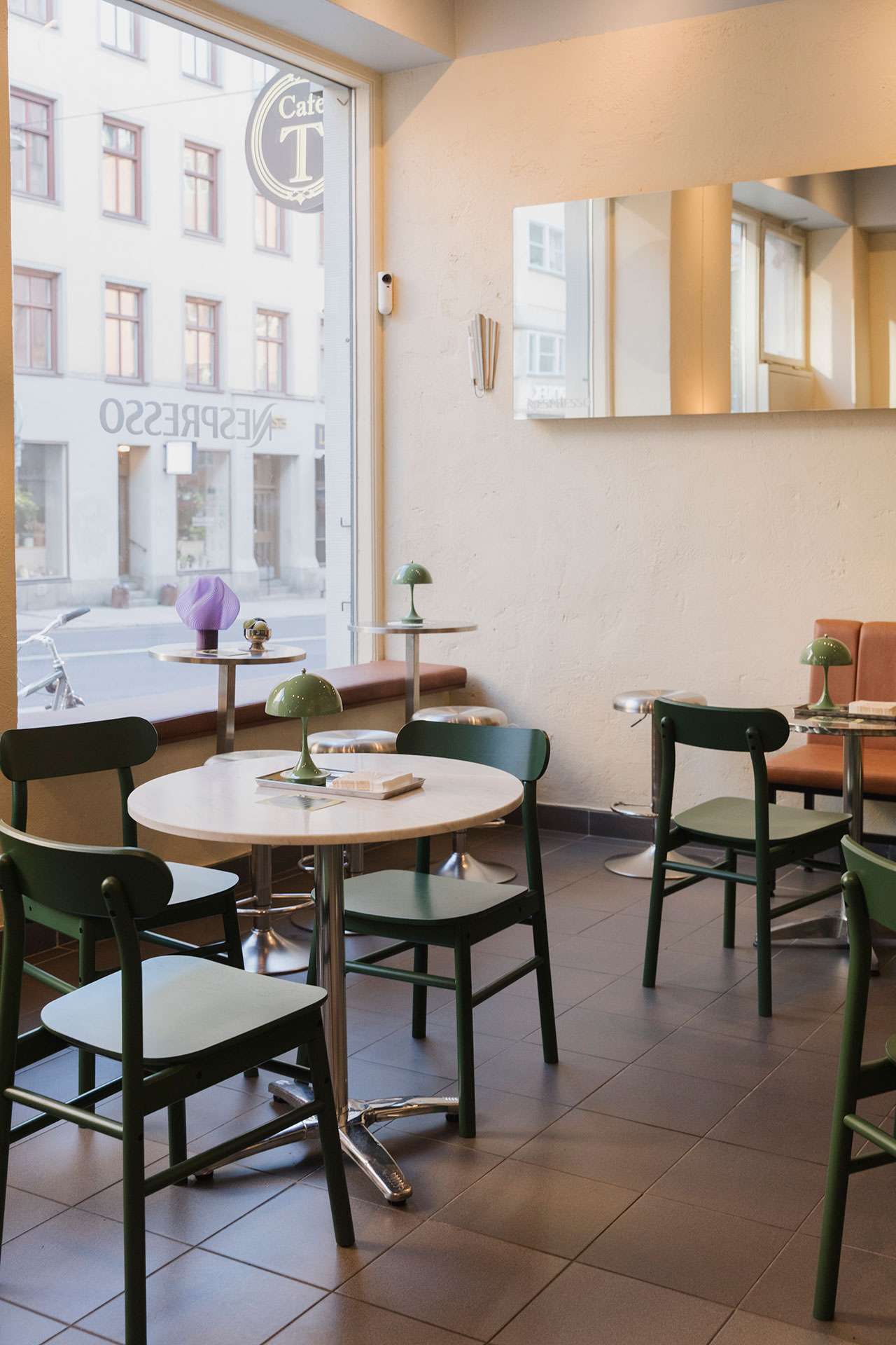 A modern café interior with round tables, dark green chairs, and small green lamps. Nespresso machines sit on the counter. Large windows reveal a street view with buildings and a person walking outside.