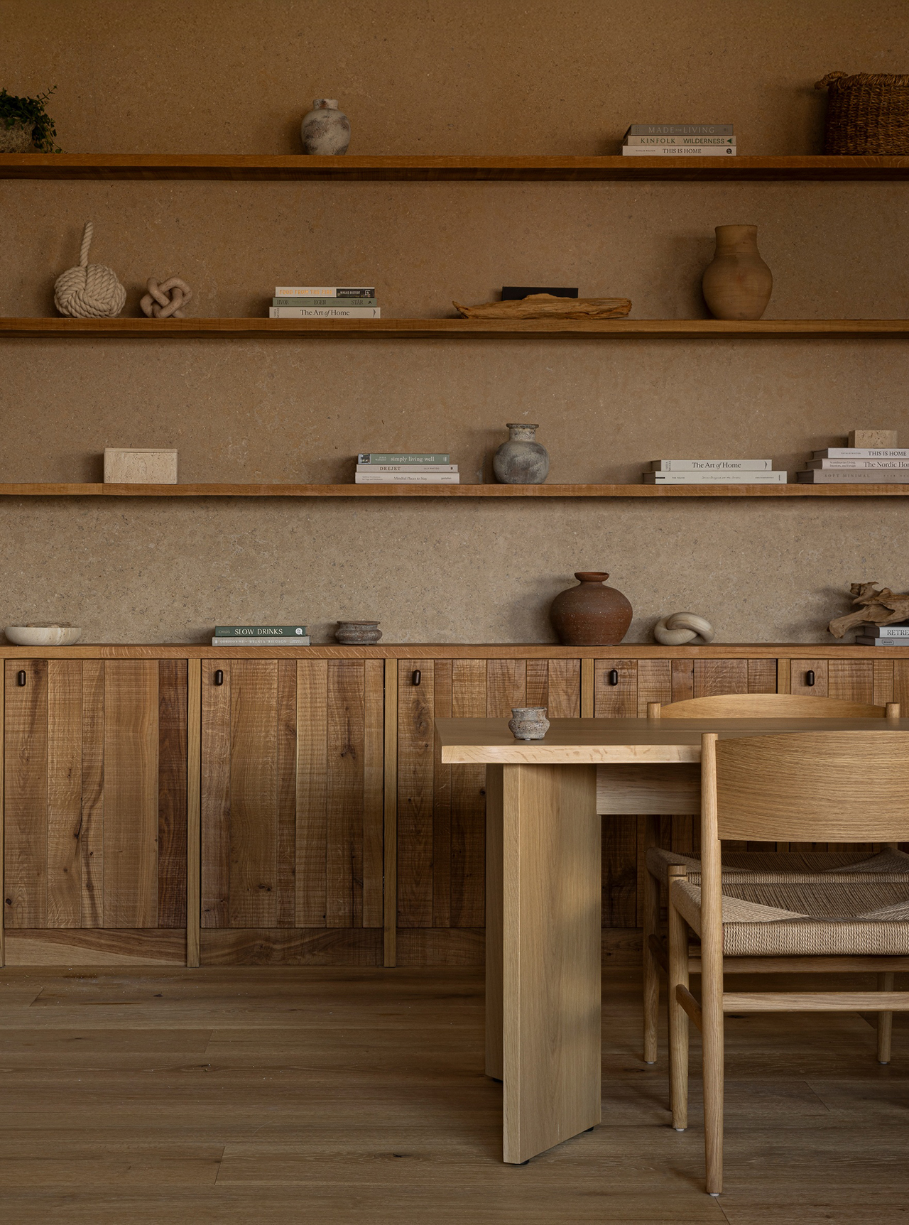 Minimalist wooden dining area inspired by Norm Architects, featuring a light wood table and chair, open shelves displaying books, ceramics, and decorative objects, with a textured neutral wall and cabinetry.