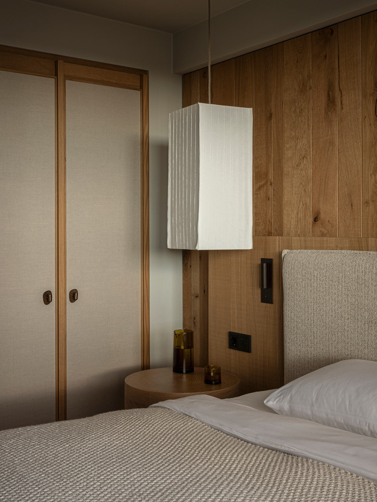 A modern bedroom by Norm Architects features a textured bed, wooden panel wall, pendant lamp, round nightstand, and double closet doors.