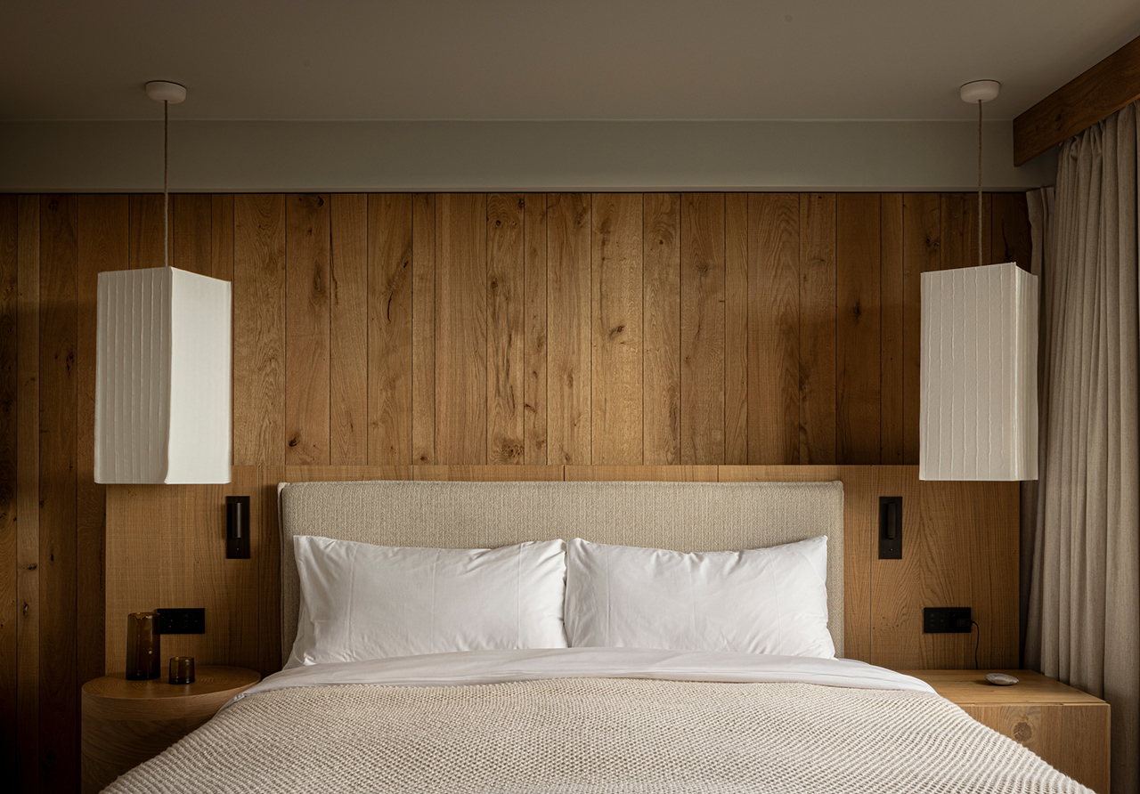 A neatly made bed with white linens and pillows is centered against a wooden paneled wall, showcasing the minimalist style of Norm Architects, and flanked by two rectangular white pendant lights and nightstands.
