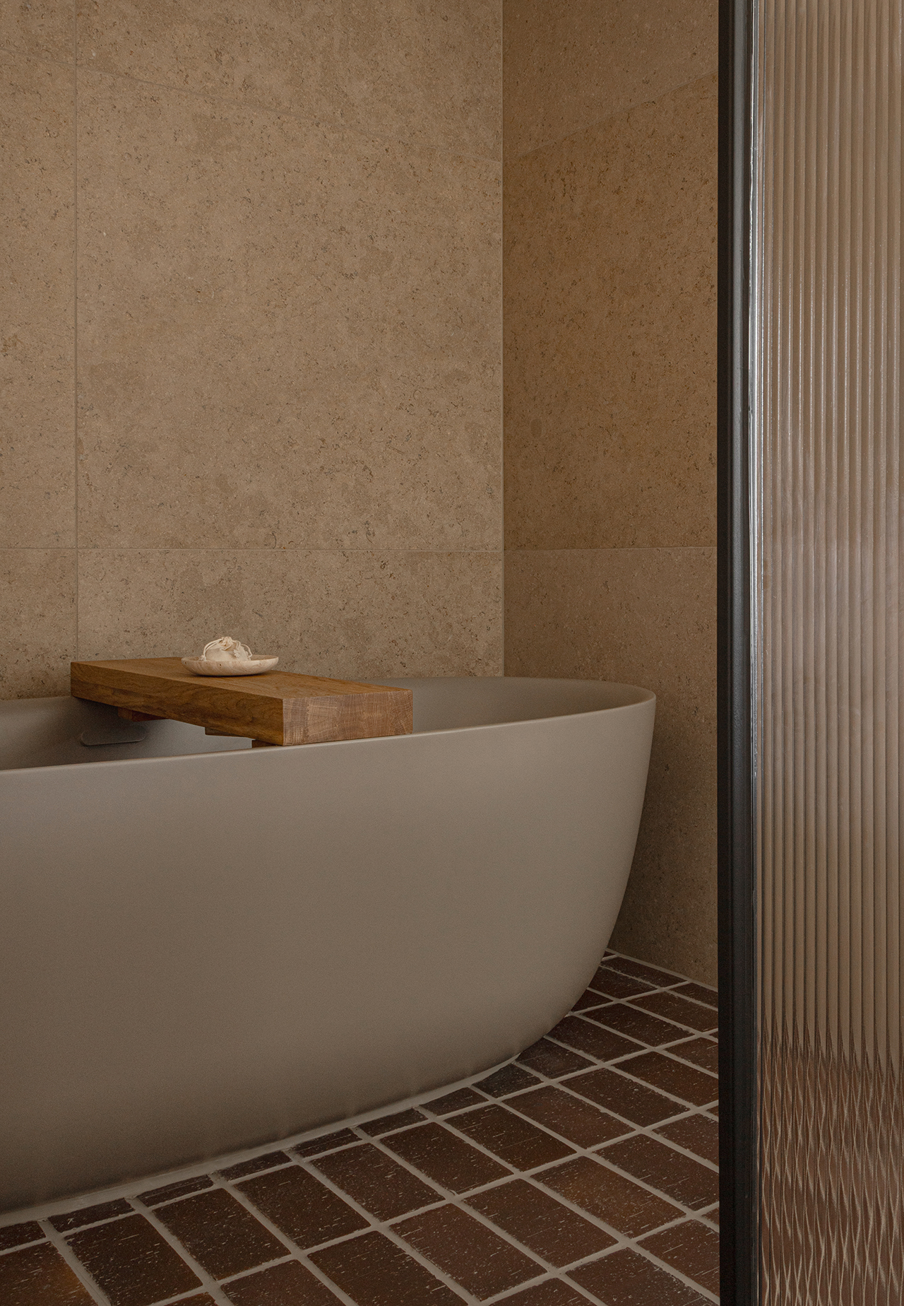 Minimalist bathroom by Norm Architects featuring a freestanding oval bathtub, wooden bath tray with a shell, beige stone walls, and a brick floor.