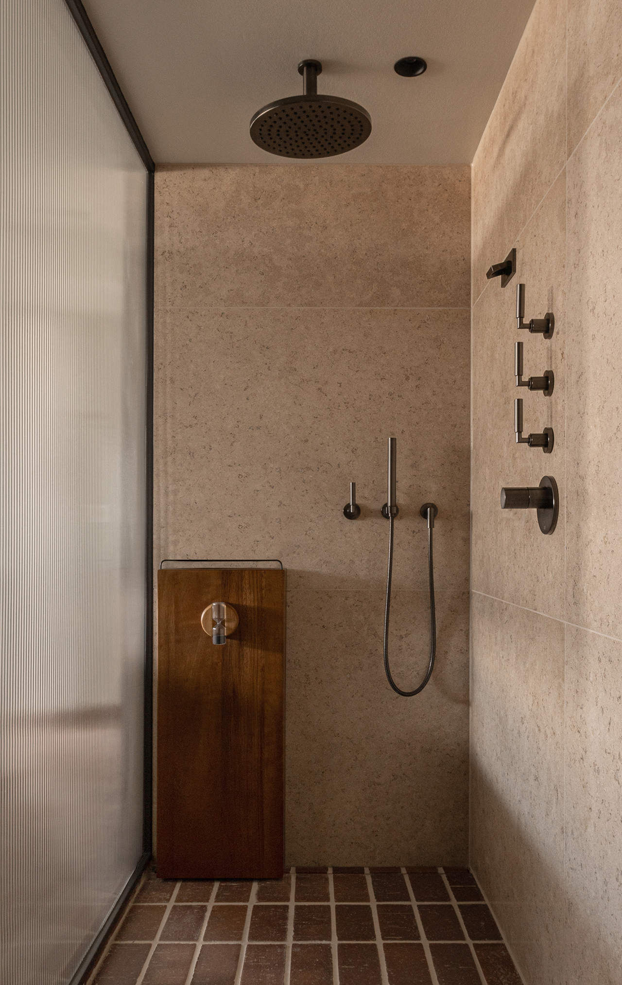 Modern walk-in shower by Norm Architects with beige stone walls, a rainfall showerhead, wall-mounted fixtures, handheld shower, and a wooden stool on a brick tile floor.