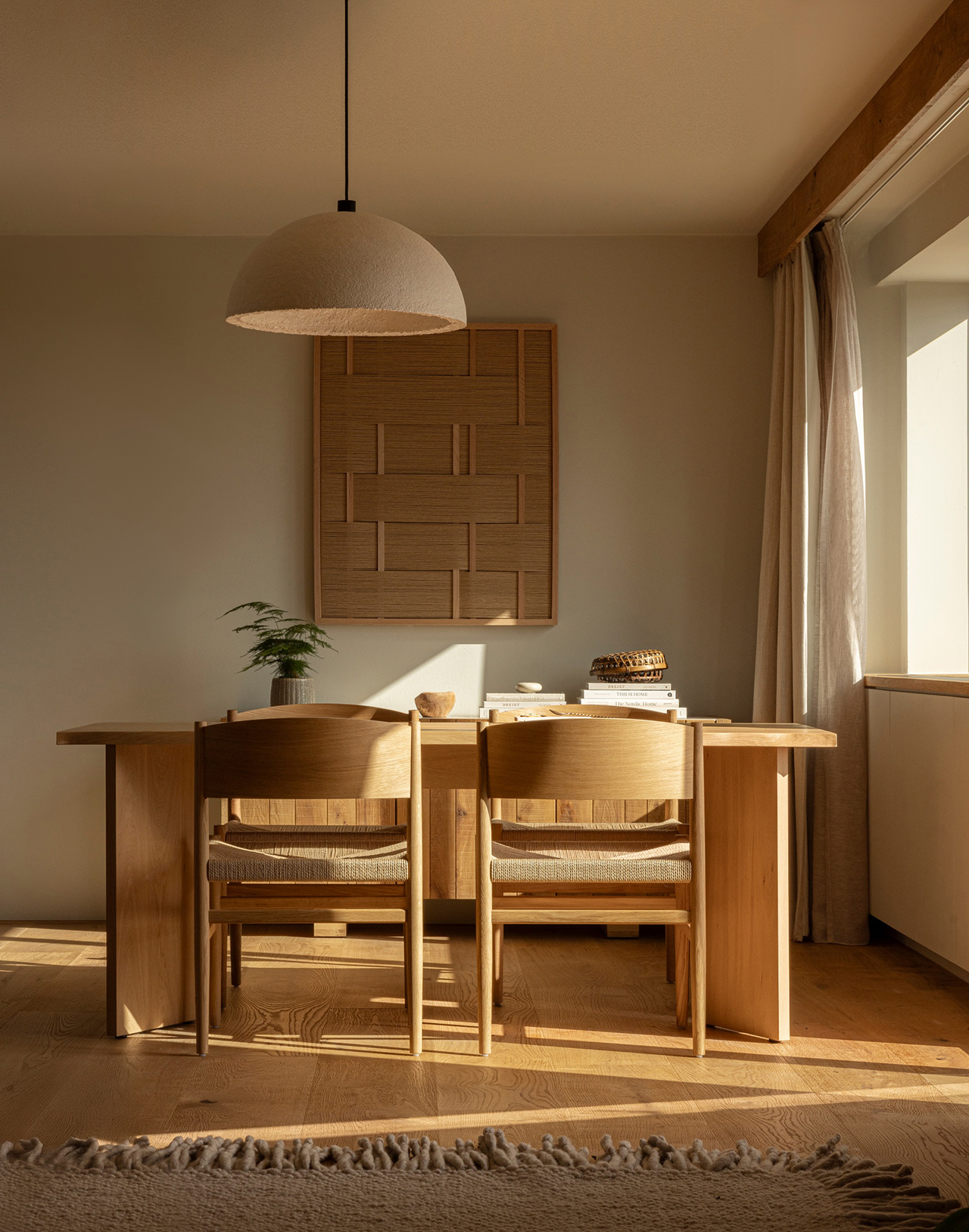 A minimalist dining area designed in the spirit of Norm Architects, featuring a wooden table, two chairs, a small plant, stacked books, and wall art, all bathed in natural sunlight from a large window.